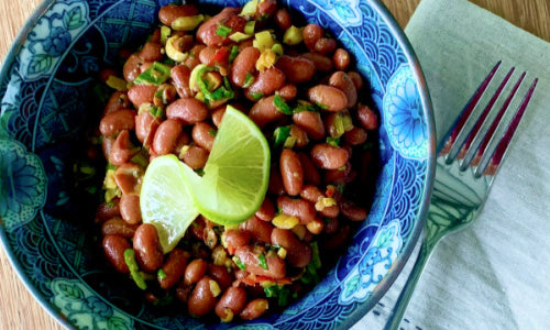 Bowl of bean salad with a slice of lime on top placed in a blue rimmed bowl beside a fork on top of a linen napkin