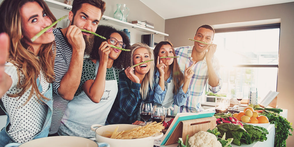 Six people at a kitchen counter taking a selfie with asparagus under their noses like mustaches 